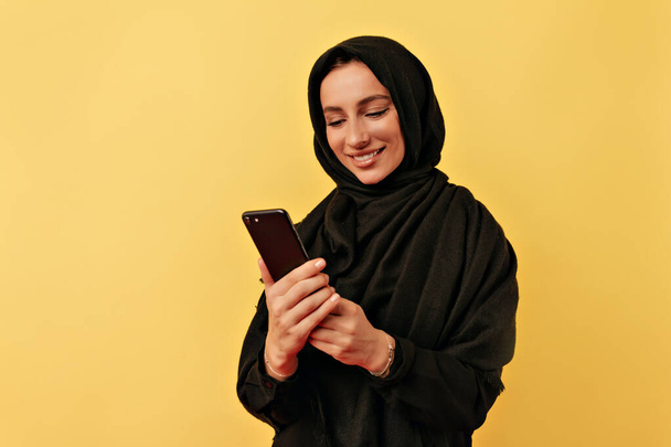 Piękny szczęśliwy uśmiechnięty Arabian kobieta ubrany czarny burki patrząc na smartphone przez odizolowany żółty tło - Zdjęcie, obraz