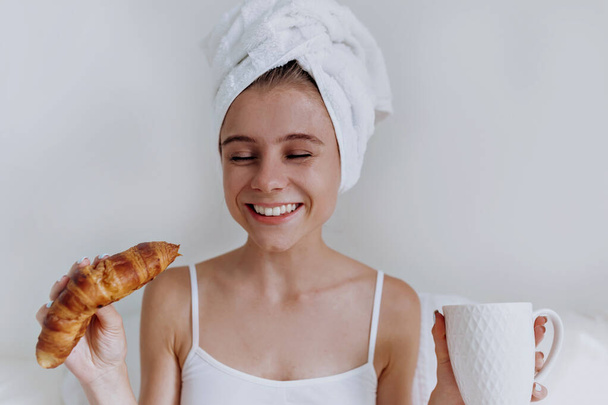 Молодая счастливая улыбающаяся привлекательная женщина с полотенцем на голове сидит в большой белой кровати и пьет кофе и ест круассан
 - Фото, изображение