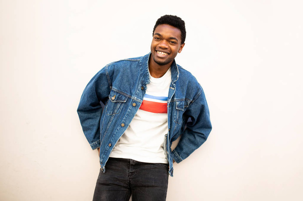 Portrait of cool african american man smiling with denim jacket leaning against white wall - Photo, Image