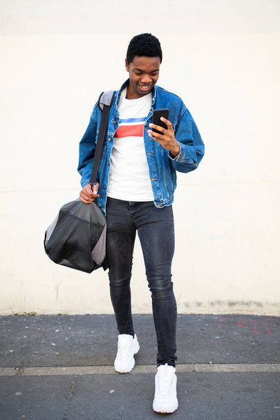 Ganzkörperporträt eines glücklichen jungen afrikanisch-amerikanischen Mannes, der mit Tasche und Handy auf der Straße läuft - Foto, Bild