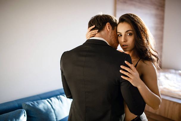 Young sexy couple in living room. Beautiful attractive young woman in black lingerie embrace man and look on camera. Businessman touch model with passion. - Valokuva, kuva