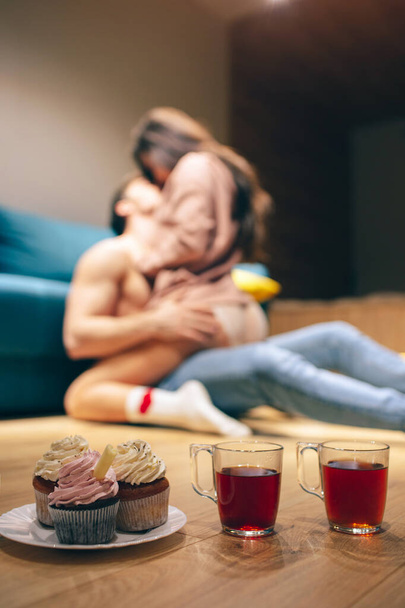 Young sexy couple have intimacy in kitchen in night. Seductive woman sit on man and kiss him. Shirtless slim guy on floor embrace model. Blurred background. Tea and pancakes on tabel. - Φωτογραφία, εικόνα