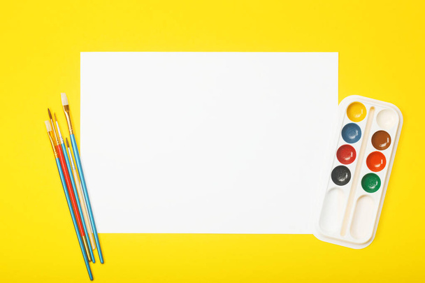 Blank paper with brushes and paints on yellow background. Artist workplace concept with copy space for your ideas. - Foto, Imagen