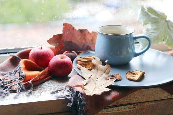 Чашка кофе на фоне окна, за которым дождь, яблоки, теплый шарф, листья, осенний сезон, концепция домашнего уюта
 - Фото, изображение
