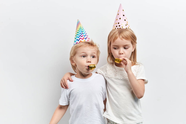 two cute children using party whistles, wearing party hats on a white background - Фото, изображение