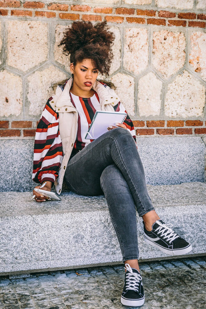 Afro vlasy brunetka studentka čte její notebook, zatímco drží tablet v druhé ruce, sedí na ulici - Fotografie, Obrázek