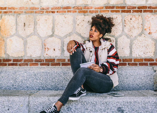 Hermosa chica morena con pelo afro está sentada en un banco de piedra, pensando en algo para escribir en su cuaderno, en un día de otoño
 - Foto, Imagen