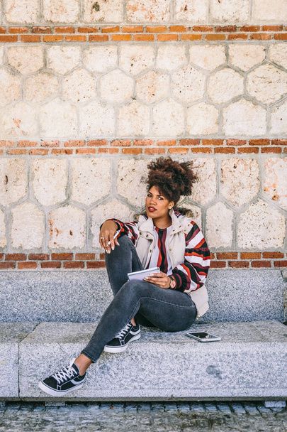 mooi brunette meisje met afro haar zitten op een stenen bank, kijkt naar de camera terwijl ze denkt iets te schrijven in haar notebook - Foto, afbeelding