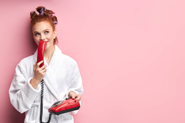 Portrait of beautiful dreamy girl wearing bathrobe standing isolated over pink background, talking on landline phone - Foto, afbeelding