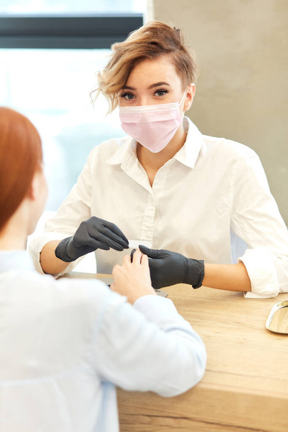 Masked master manicurist looking at camera while working with clients nails at beauty salon - 写真・画像