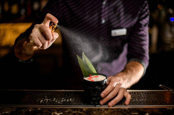 Bartender spaying on a black glass with cocktail decorated with a green tropical leaf, dried rose bud and essence drops - Фото, изображение