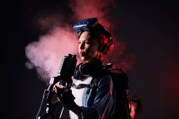 Young caucasian woman in special clothes wearing virtual reality headset goggles holding gun - Zdjęcie, obraz