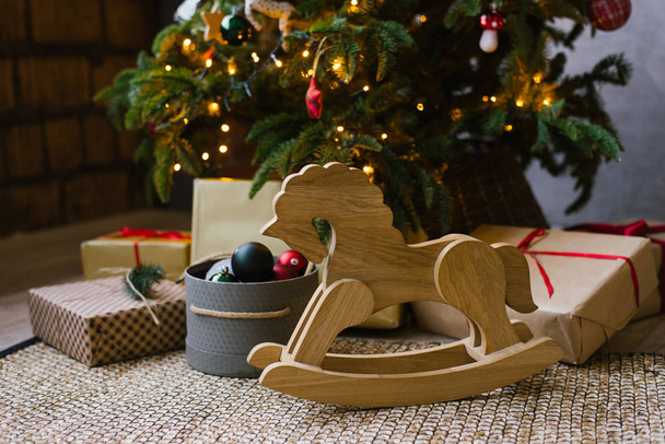 A wooden rocking horse for children stands next to Christmas gifts under a Christmas tree with lights - Foto, Imagen