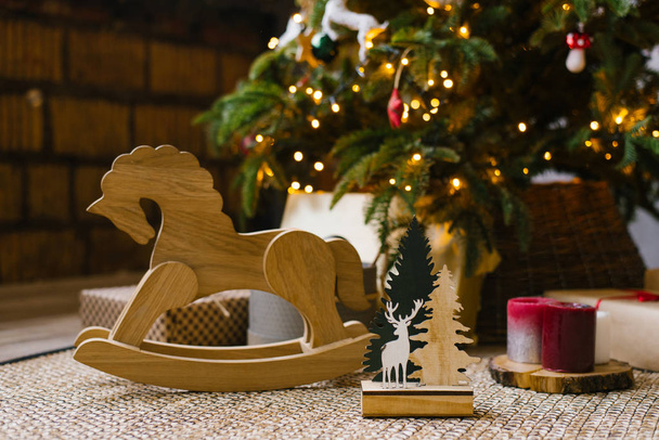 Een houten schommelpaard voor kinderen staat naast kerstcadeaus onder een kerstboom met lichtjes - Foto, afbeelding