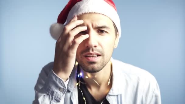 Closeup footage of young attractive man in santa claus red hat after celebration or long night looking at camera mirror and touching his face, recognizing himself, preen. Background isolated on blue - Video