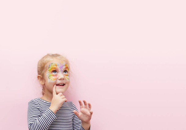 Pretty exciting smiling blond little child girl of 3 years with a bright face painting on pink background. Fun emotions, happy childhood - Foto, imagen