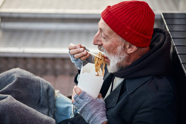 Old-aged vagabond sitting on walking street and hungrily eating - Foto, Imagem