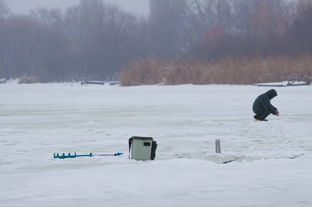 zimní rybaření na malém, přírodním, zamrzlém jezeře, pokrytém sněhem, ledovcem, plastovou krabicí, pruty a termoskou, figurka rybáře v teplých šatech sedící a snažící se ulovit ryby, hobby volnočasová image - Fotografie, Obrázek