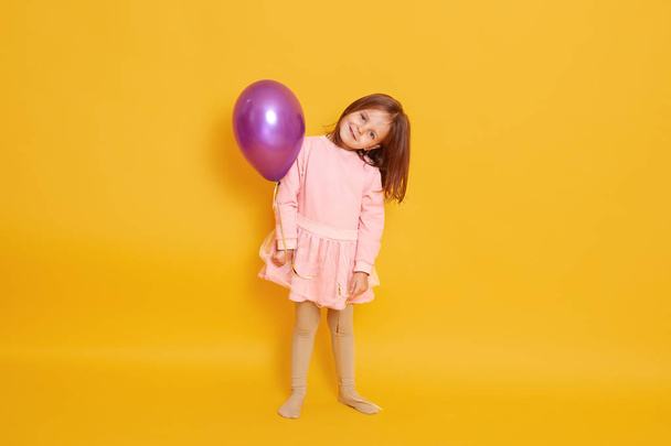 Horizontal shot of cute little girl holds balloon isolated over yellow background, female child wears pink dress, kid with dark hair, looks smiling directly at camera, has birthday. Childhood concept. - Photo, Image
