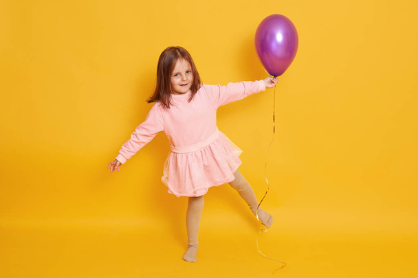Studio shot of beautiful little girl wih bright purple balloon in hands, child standing on one hand isolated over yellow studio background, female kid wearing rosy dress, celebrating her birthday. - Fotografie, Obrázek