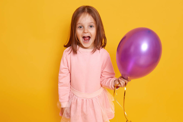 Portrait of surprised little girl with dark staright hair standing over yellow studio background beautiful clothes, holding purple ballon in hands and looking at camera joyfully. Holiday concept. - Photo, image