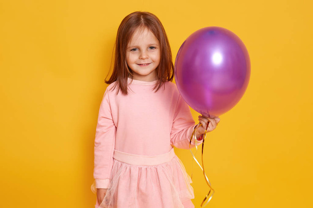 Close up portrait of sweet nice girl with purple baloon in hands, posing isolated over yellow background, adorable female child wearing pink dress, looks smiling directly at camera. Party concept. - Fotografie, Obrázek
