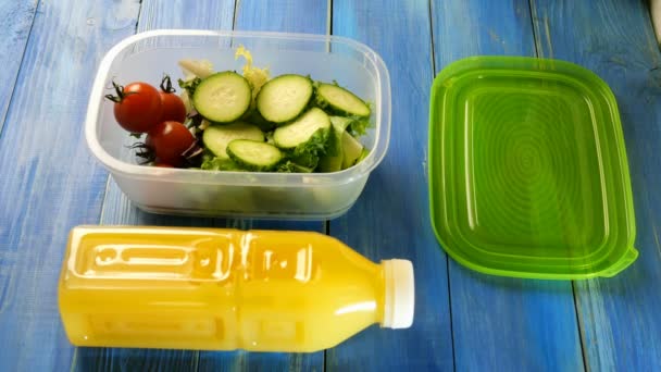 Human hands pour with cooking oil fresh salad, cucumber and cherry tomatoes in plastic lunch box. Nearby is plastic bottle of orange juice. Healthy vegetarian food concept. Top view. Close-up. - 映像、動画
