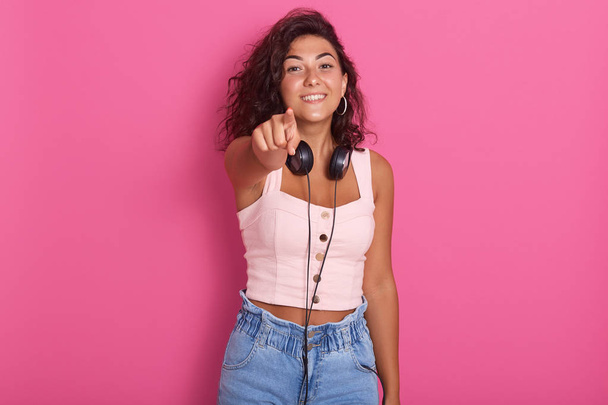 Close up portrait of pretty girl isolated over pink studio background, poiting with her index finger to camera, charming female wearing rose top and jeans, hacing dark hair, girl expressing happyness. - Φωτογραφία, εικόνα