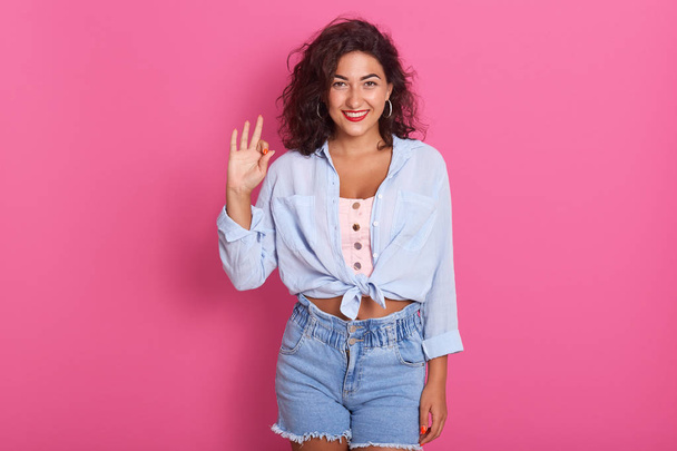 Close up portrait of happy woman showing ok sign with her hand, female wearing blue shirt and jeans shirt, lady with dark wavy hair posing isolated over pink background. Body language concept. - Foto, Bild