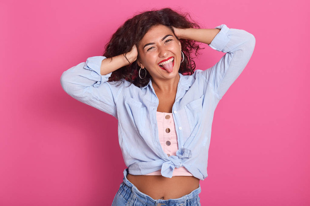 Portrait of lovely girl with dark wavy hair,showing her tongue, looking directly at camera isolated over pink background, female wearing blue shirt, shows bare belly, keeps hands on head, having fun. - Foto, Imagen