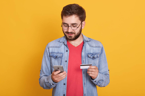 Studio shot of young european bearded man holding credit card and smartphone, standing with calm facial expression over yellow background, spending money from his account. Online shopping concept. - Foto, immagini