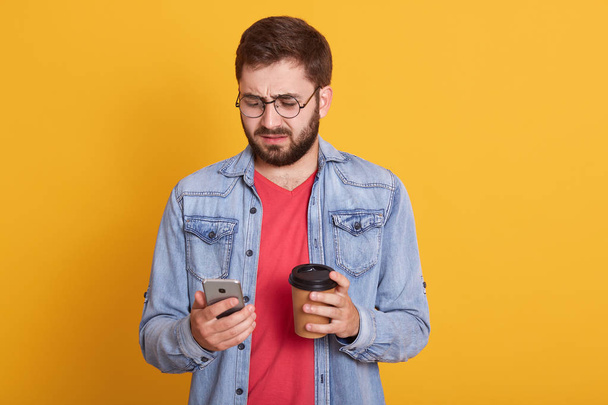 Image of handsome bearded man wearing eyewear,casual red t shirt and stylish denim jacket, holding smartphone and credit card isolated on yellow background. Online shopping and banking concept. - 写真・画像