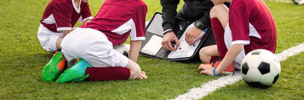 Coach coaching soccer kids soccer team. Youth sports coach using tactics board. Trainer explaining match strategy. Sports soccer education. Three junior players sitting with coach on grass pitch - Photo, Image