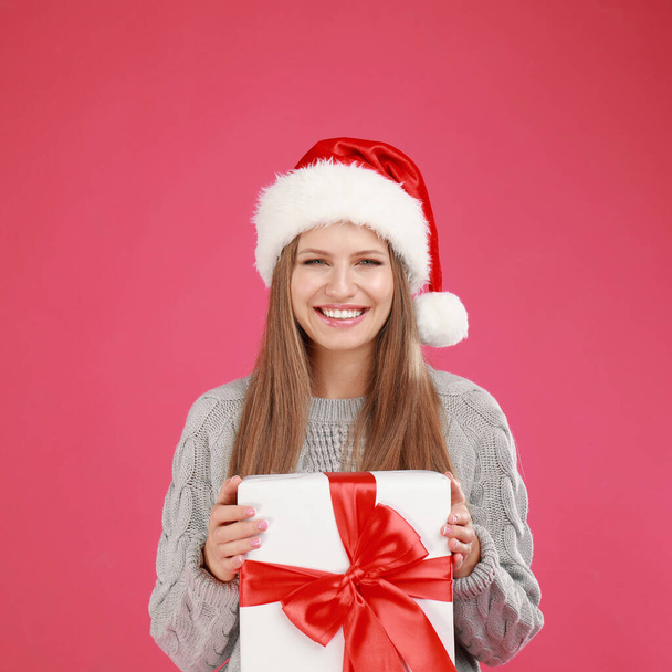 Happy young woman in Santa hat with Christmas gift on pink background - Фото, зображення
