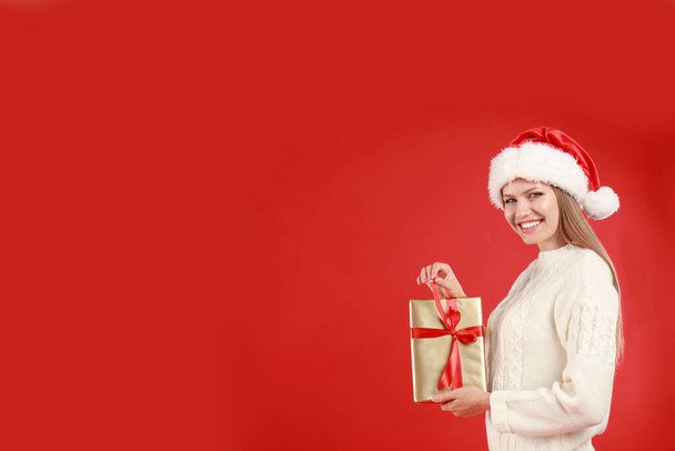 Happy young woman in Santa hat with Christmas gift on red background. Space for text - Foto, Imagem