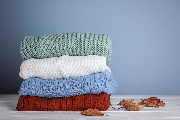 Stack of folded knitted sweaters and autumn leaves on white wooden table against grey background. Space for text - Φωτογραφία, εικόνα