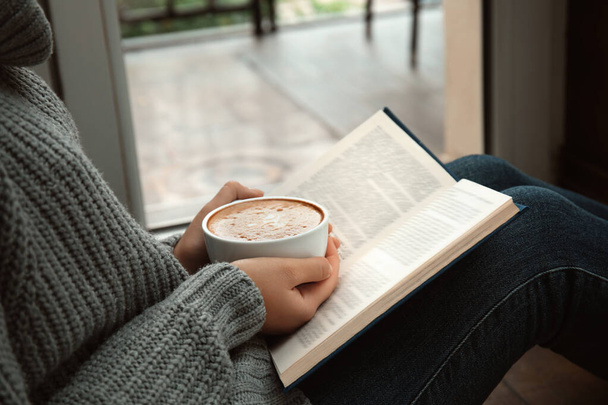 Mulher com xícara de livro de leitura de café perto da janela dentro de casa, close-up
 - Foto, Imagem