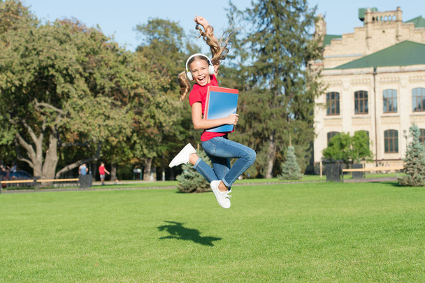 Starting school in good mood. Happy schoolgirl jump to music on green grass. Active little child in headphones hold school books. Back to school. School holidays. Summer vacation. Primary education - Foto, imagen