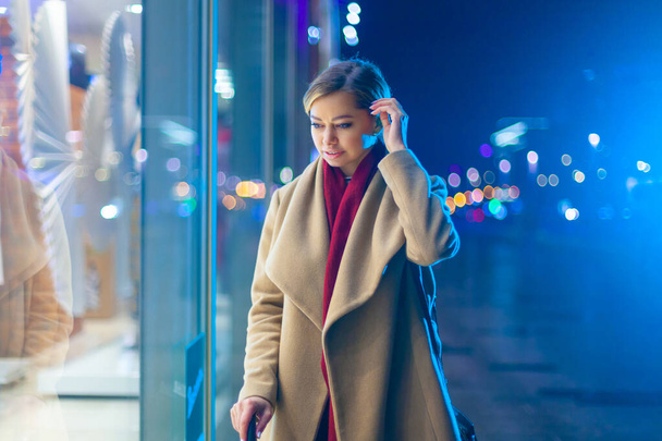 Christmas. A young beautiful woman in the late evening looks at the glowing window and chooses a gift. Colorful, magical lighting, beautiful glare. Cheerful girl wondered at the window, the agony of - Foto, Imagem