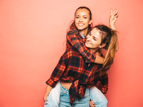 Two young beautiful smiling brunette hipster girls in trendy similar checkered shirt and jeans clothes.Sexy carefree women posing near pink wall in studio.Positive model sitting on her friend back - Photo, image