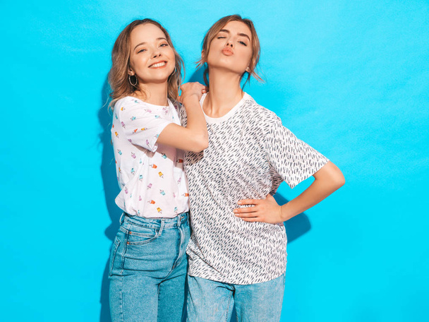 Two young beautiful smiling blond hipster girls in trendy summer colorful T-shirt clothes. Sexy carefree women posing near blue wall. Positive models having fun.Winks and makes duck face - Photo, image