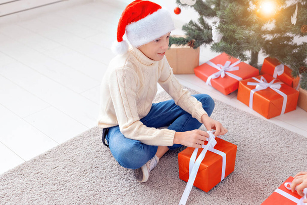 Διακοπές, Χριστούγεννα, παιδική ηλικία και άνθρωποι έννοια - χαμογελαστός χαρούμενος έφηβος αγόρι στο καπέλο Σάντα ανοίγει κουτί δώρου πάνω από το φόντο χριστουγεννιάτικο δέντρο - Φωτογραφία, εικόνα