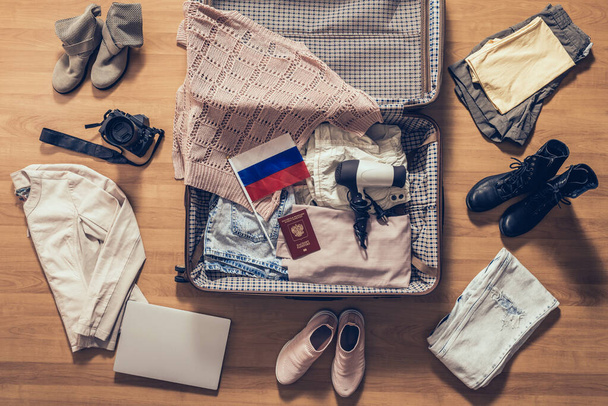 Vestiti da donna, laptop, macchina fotografica, passaporto russo e bandiera distesi sul pavimento del parquet vicino e nella valigia aperta. Concetto di viaggio
 - Foto, immagini