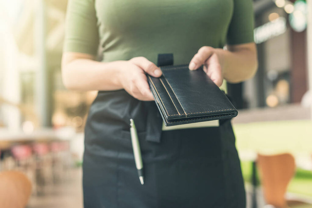 Cropped image of woman waitress in apron hold out bill folder to cafe visitor of cafe or restaurant. - Foto, immagini