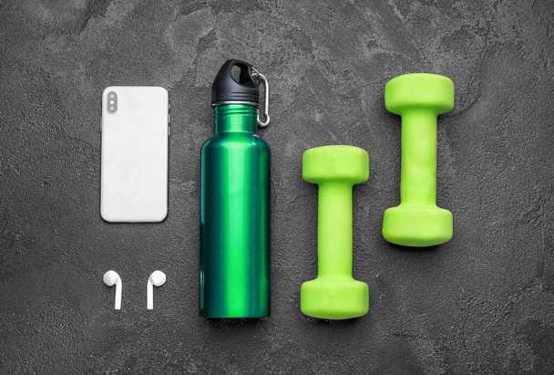 Sports water bottle, earphones, mobile phone and dumbbells on dark background - Фото, изображение