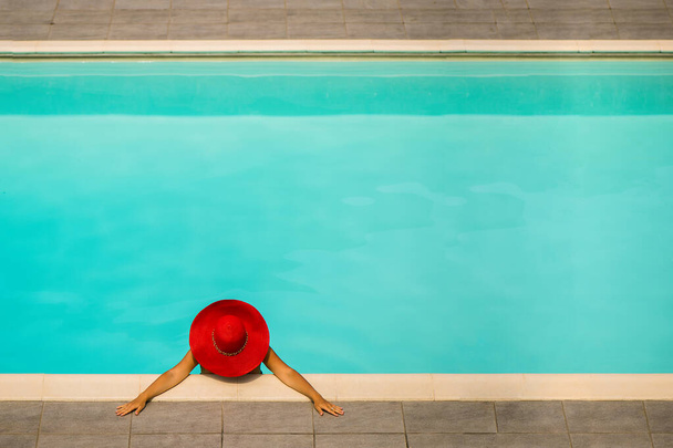 Όμορφο κορίτσι με μαγιό και κόκκινο καπέλο σε μια γαλάζια πισίνα. - Φωτογραφία, εικόνα