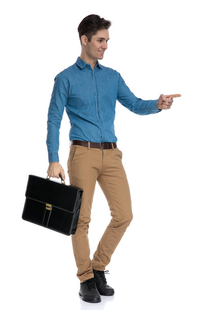 inteligentní ležérní muž držící kufr a ukazující prstem - Fotografie, Obrázek