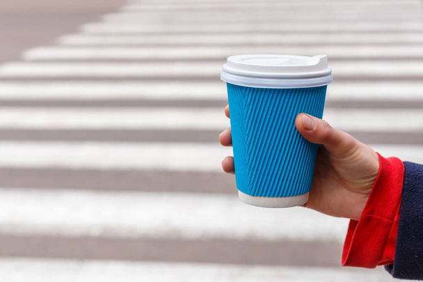 Κοντινό πλάνο γυναίκα κρατώντας ένα μπλε χάρτινο φλιτζάνι καφέ στο χέρι σε φόντο διασταύρωση ζέβρας. - Φωτογραφία, εικόνα