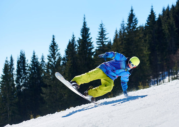 Snowboarder man in goggles and helmet riding snowboard fast down steep snowy mountain slope on background of blue sky and spruce trees on sunny winter day - Photo, Image