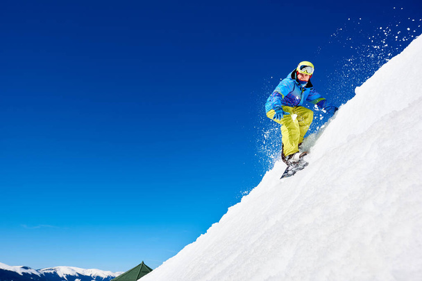 Snowboarder man in goggles and helmet riding snowboard fast down steep snowy mountain slope on background of blue sky and spruce trees on sunny winter day - Photo, Image